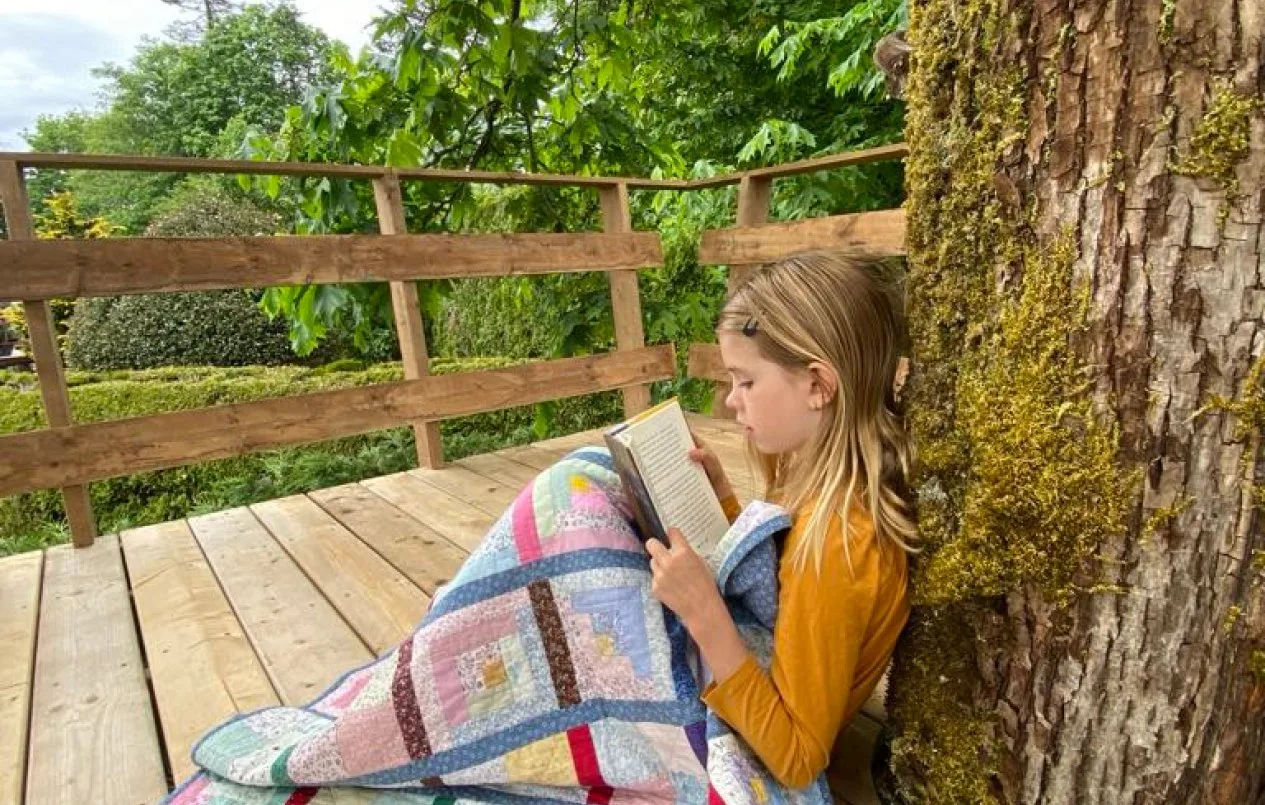 Child reading a book under a quilt on an outdoor deck